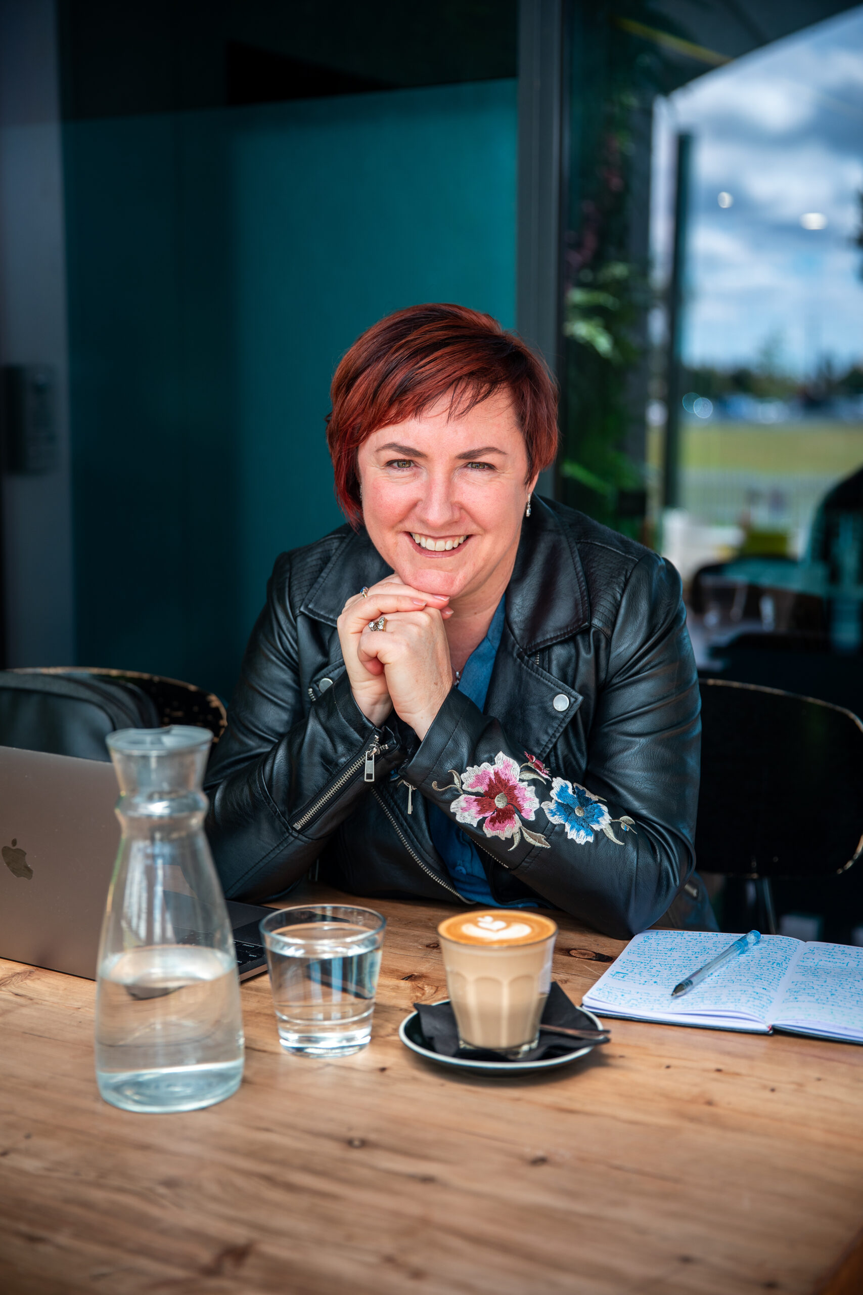Kerrie sits in a cafe at a wooden table with her chin resting on her hands. She wears a black leather jacket with floral embroidery on the left sleeve. On the table in front of her is a laptop, pen on top of a journal, cup of coffee and a carafe and glass of water.