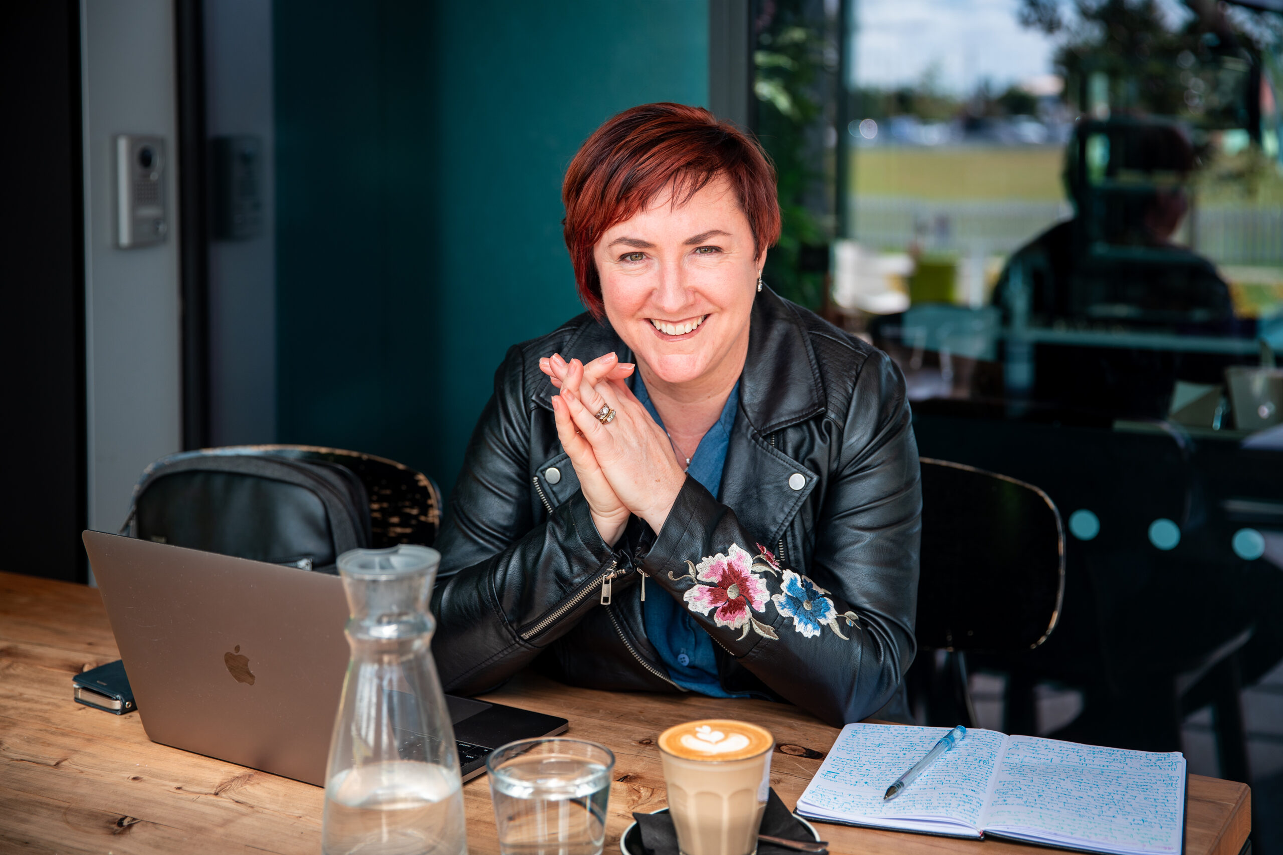 Kerrie sits in a cafe at a wooden table with her hands together. She wears a black leather jacket with floral embroidery on the left sleeve. On the table in front of her is a laptop, pen on top of a journal, cup of coffee and a carafe and glass of water.